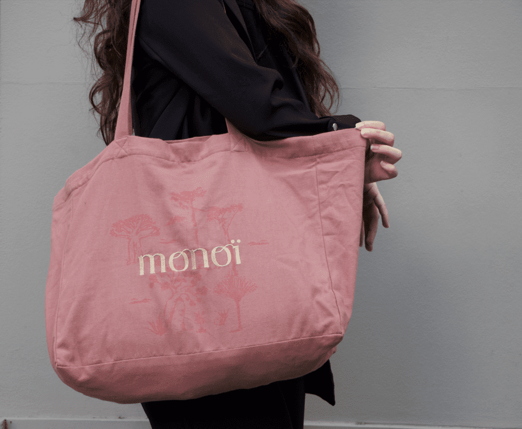 Une personne en tenue sombre porte un tote bag rose avec le mot « monoï » et un dessin illustré dessus.