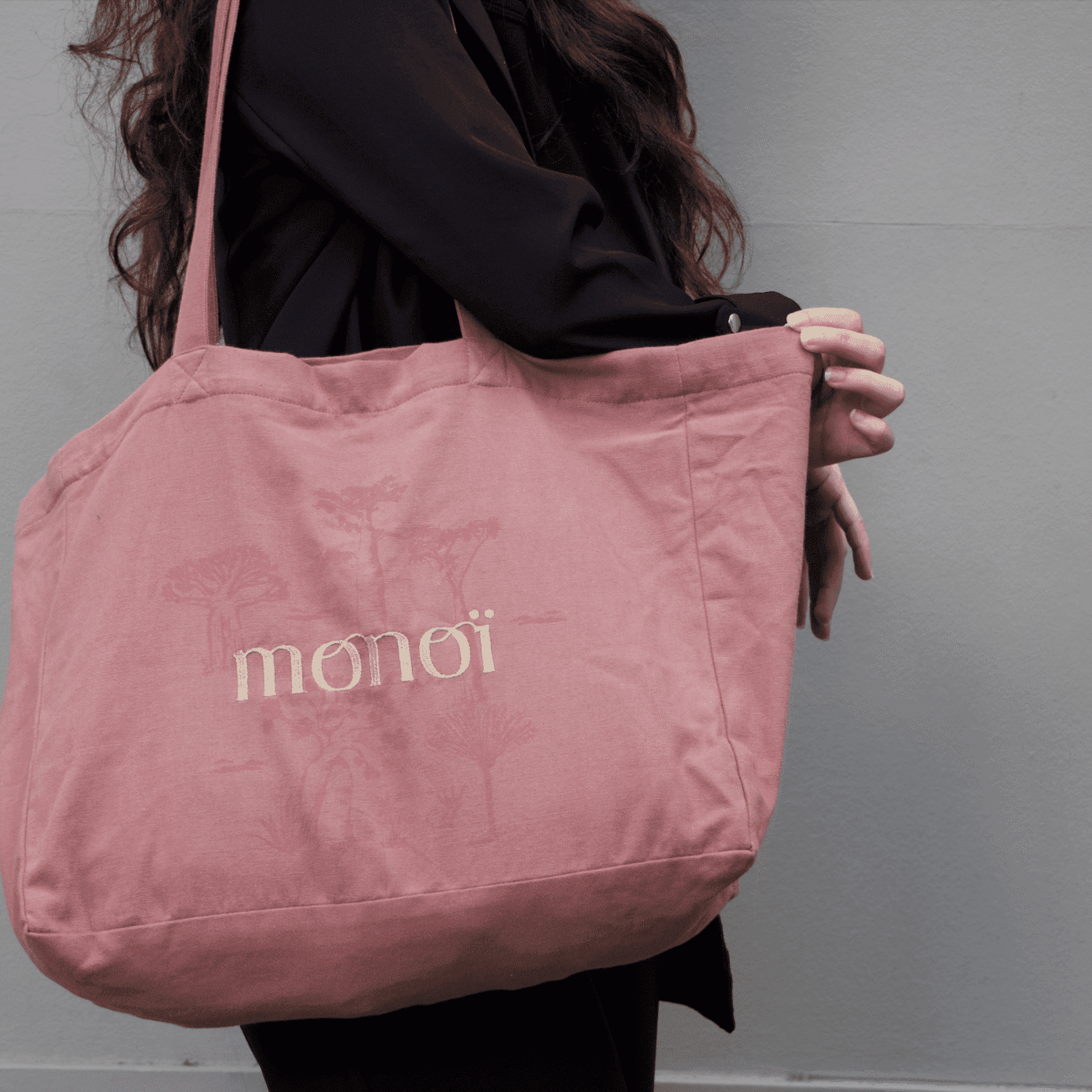 Une personne en tenue sombre porte un tote bag rose avec le mot « monoï » et un dessin illustré dessus.