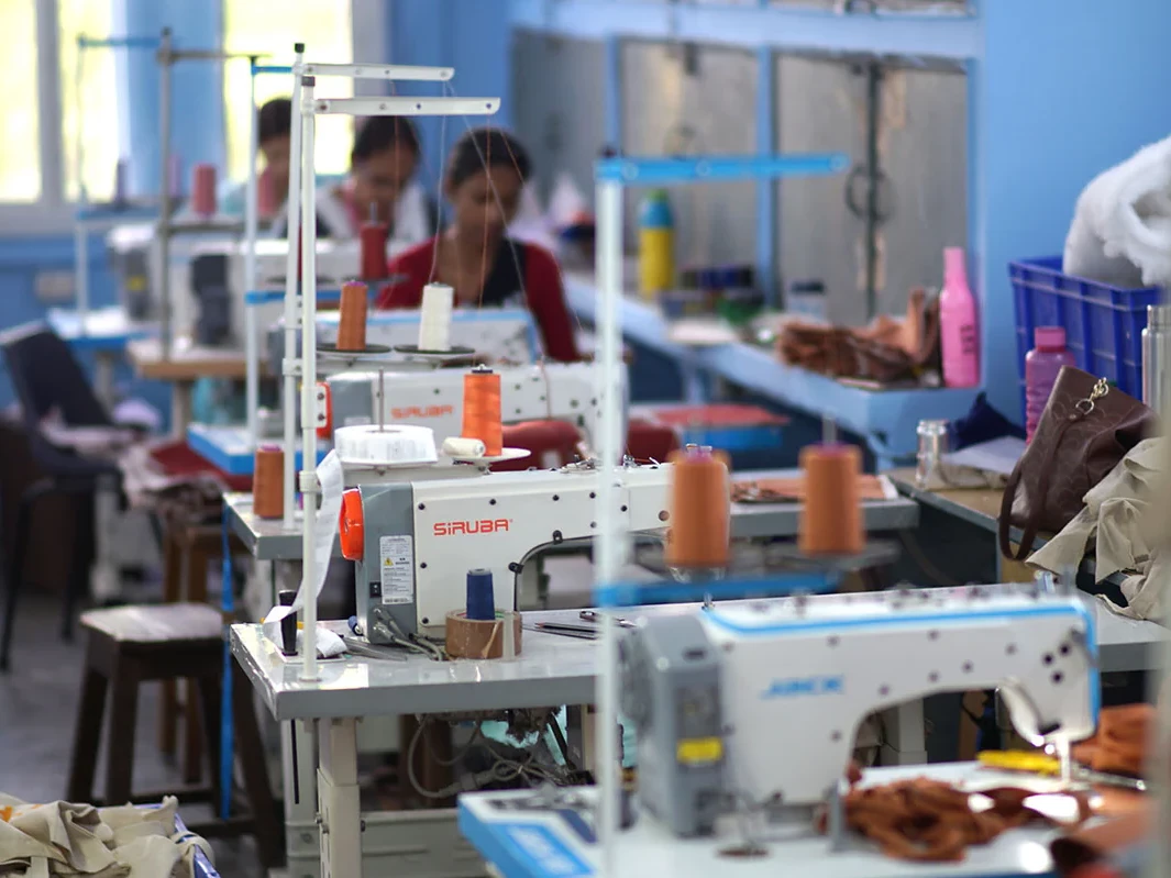 Image d'une usine de confection o&ugrave; des gens travaillent sur des machines &agrave; coudre dans une pi&egrave;ce bien &eacute;clair&eacute;e avec des &eacute;tag&egrave;res contenant diverses fournitures.