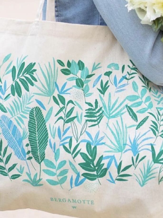 Une personne tient un sac fourre-tout beige avec un motif de feuilles vertes et bleues et le mot « Bergamotte » imprimé en bas, ainsi qu'un bouquet de fleurs blanches.