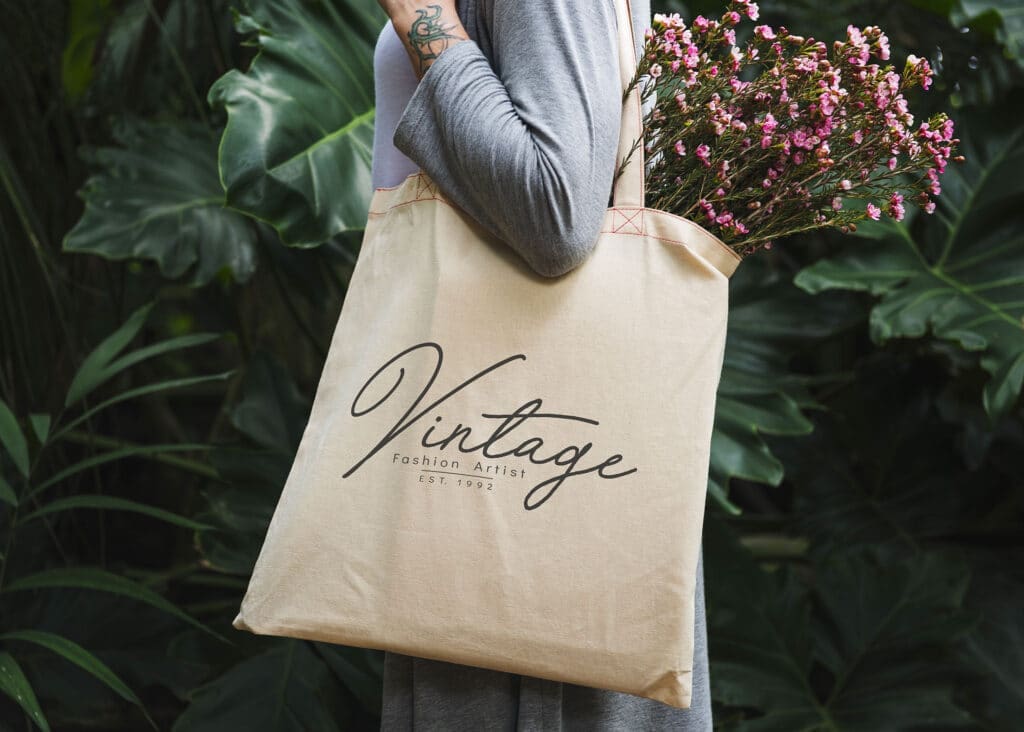 Personne tenant un sac fourre-tout beige sur lequel est imprimé le mot « Vintage » et portant des fleurs roses, debout devant un feuillage vert.