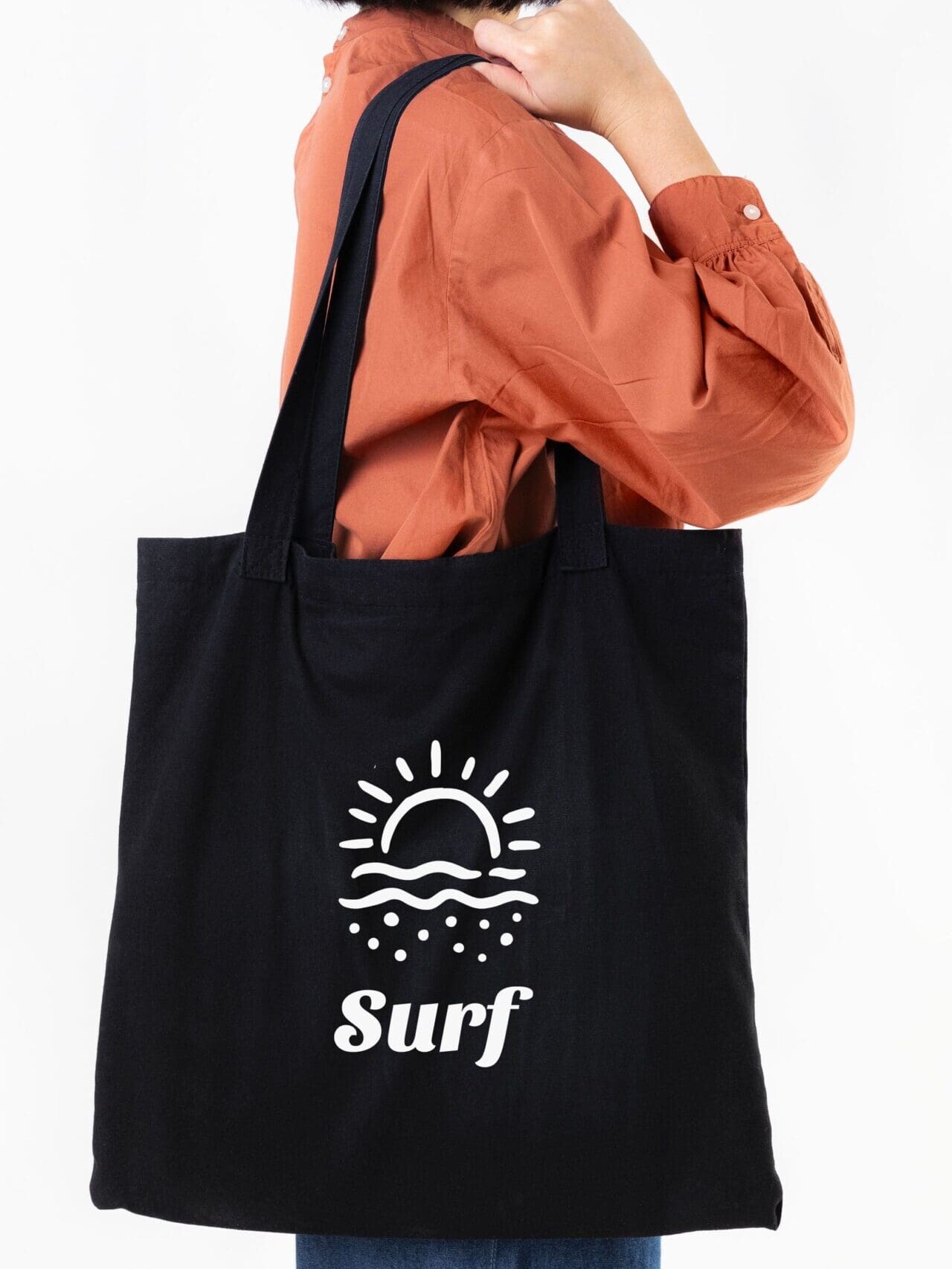 Personne tenant un sac fourre-tout noir avec un motif de surf blanc, portant une chemise orange et un jean bleu, sur un fond blanc uni.