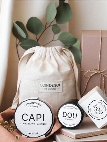 Une collection de produits de soin, dont un shampoing et une crème déodorante, avec un sac à cordon étiqueté « Soin de Soi » et un coffret cadeau marron, présenté avec des feuilles vertes et un rideau blanc en toile de fond.