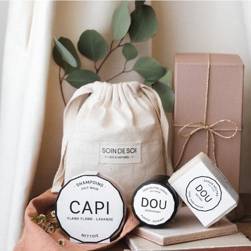 Une collection de produits de soin, dont un shampoing et une crème déodorante, avec un sac à cordon étiqueté « Soin de Soi » et un coffret cadeau marron, présenté avec des feuilles vertes et un rideau blanc en toile de fond.