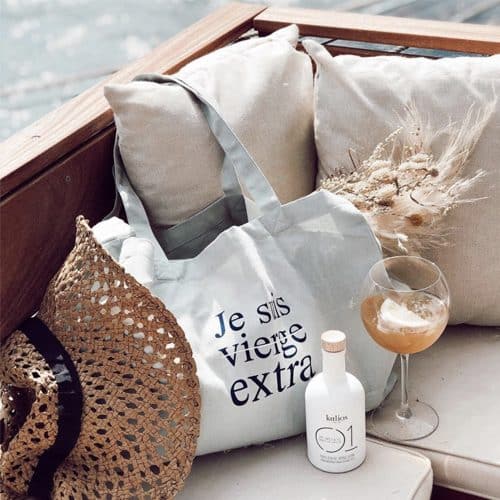 Un sac en tissu avec les mots « Je suis vierge extra » est placé sur un canapé aux côtés d'un chapeau de paille, d'une bouteille de lotion étiquetée « 01 » et d'un verre rempli d'une boisson brune.