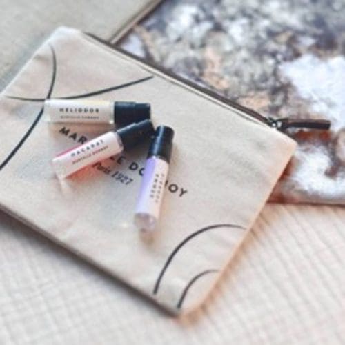 Trois petits flacons d'échantillons de parfum sont présentés sur une pochette beige zippée avec du texte noir, placée sur une surface texturée.