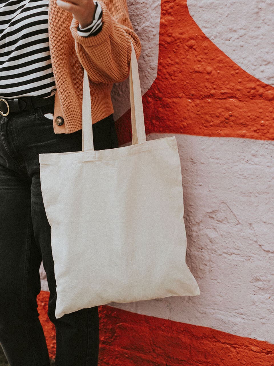 Personne portant une chemise rayée, un pull orange et un pantalon noir, tenant un sac fourre-tout beige, debout devant une fresque colorée aux motifs rouges et roses.