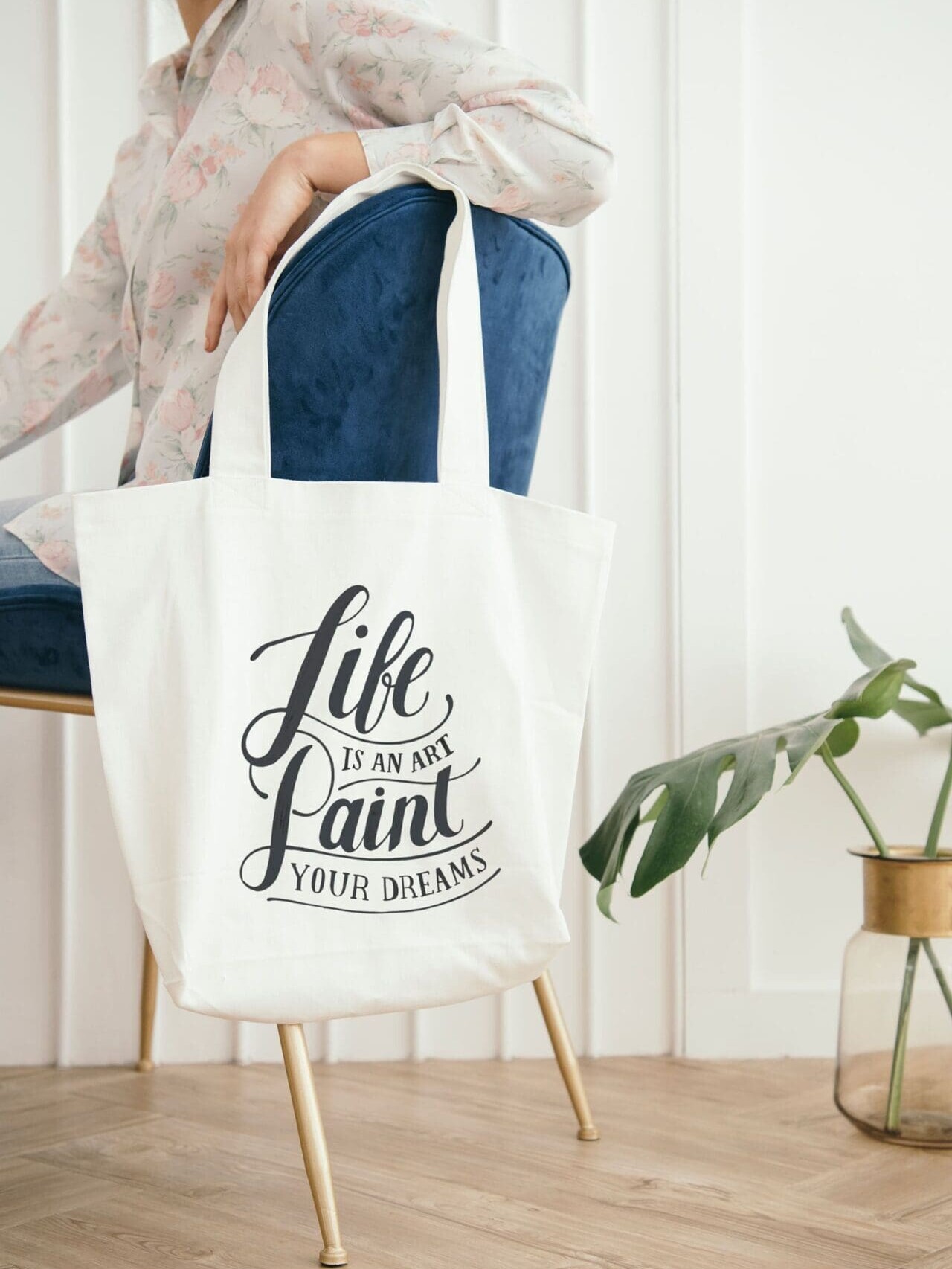 Une personne est assise sur une chaise bleue, tenant un sac fourre-tout blanc avec le texte « La vie est un art, peignez vos rêves », tandis qu'un vase de plantes décoré se trouve sur le sol à proximité.