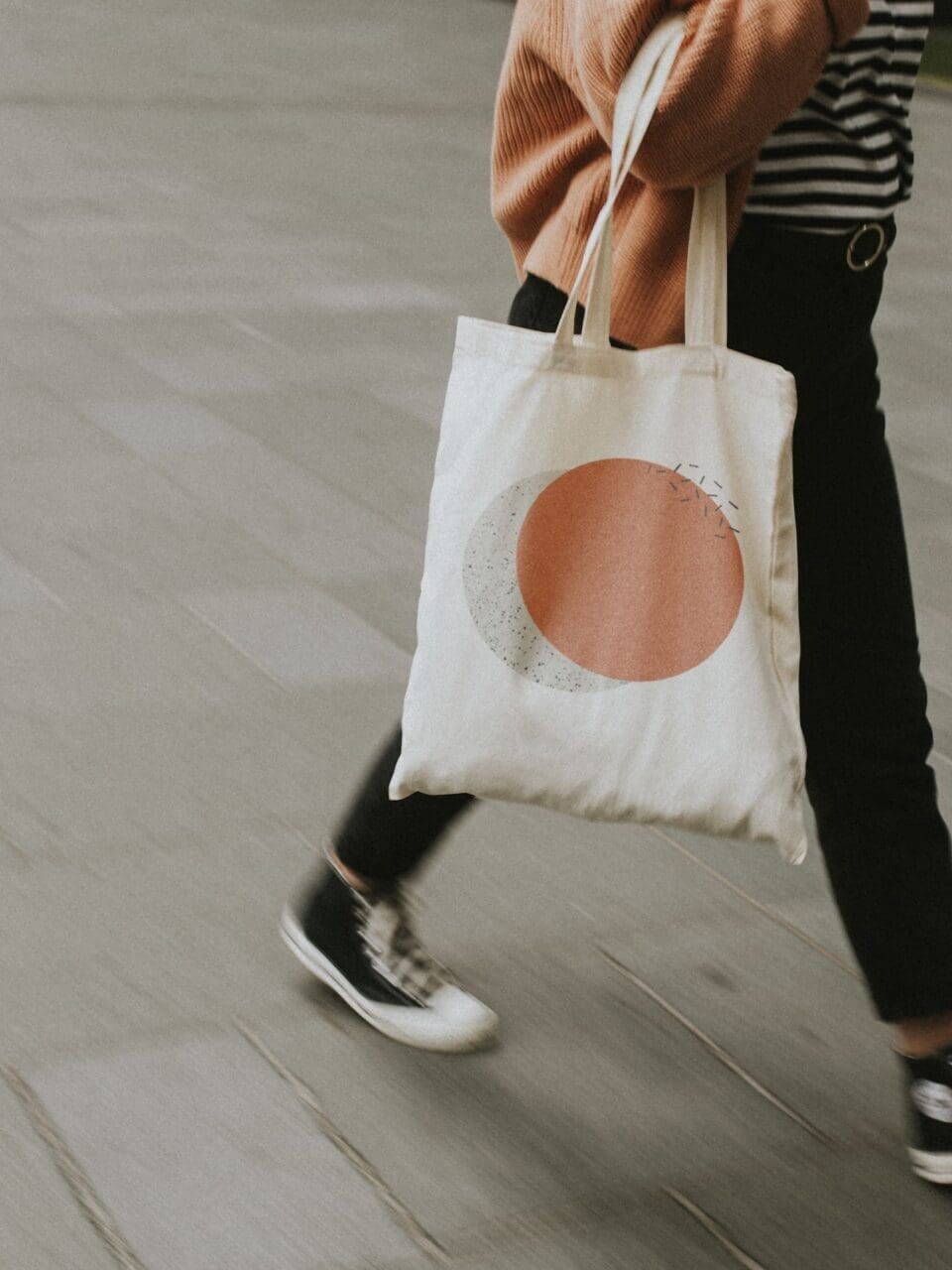 Personne marchant sur un trottoir carrelé gris portant un sac fourre-tout blanc avec un motif circulaire rose et gris.