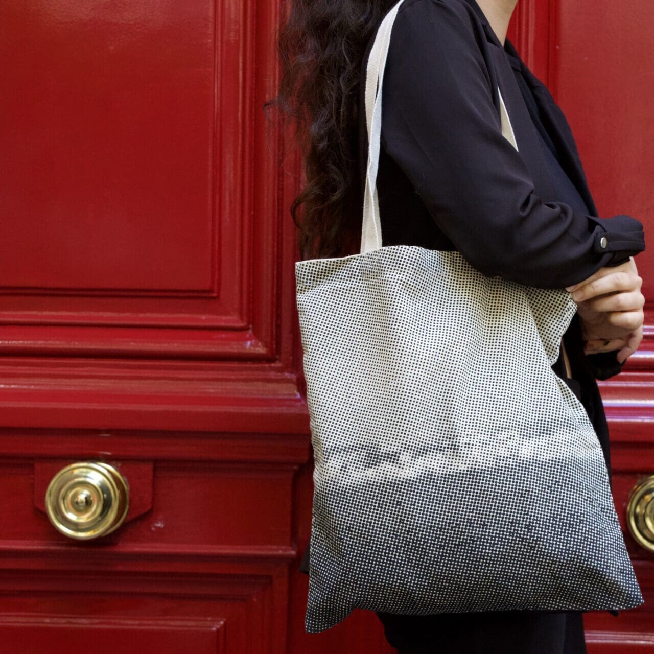 Une personne aux longs cheveux noirs, vêtue de noir, tient à deux mains un sac fourre-tout blanc et noir devant une porte rouge aux poignées dorées.