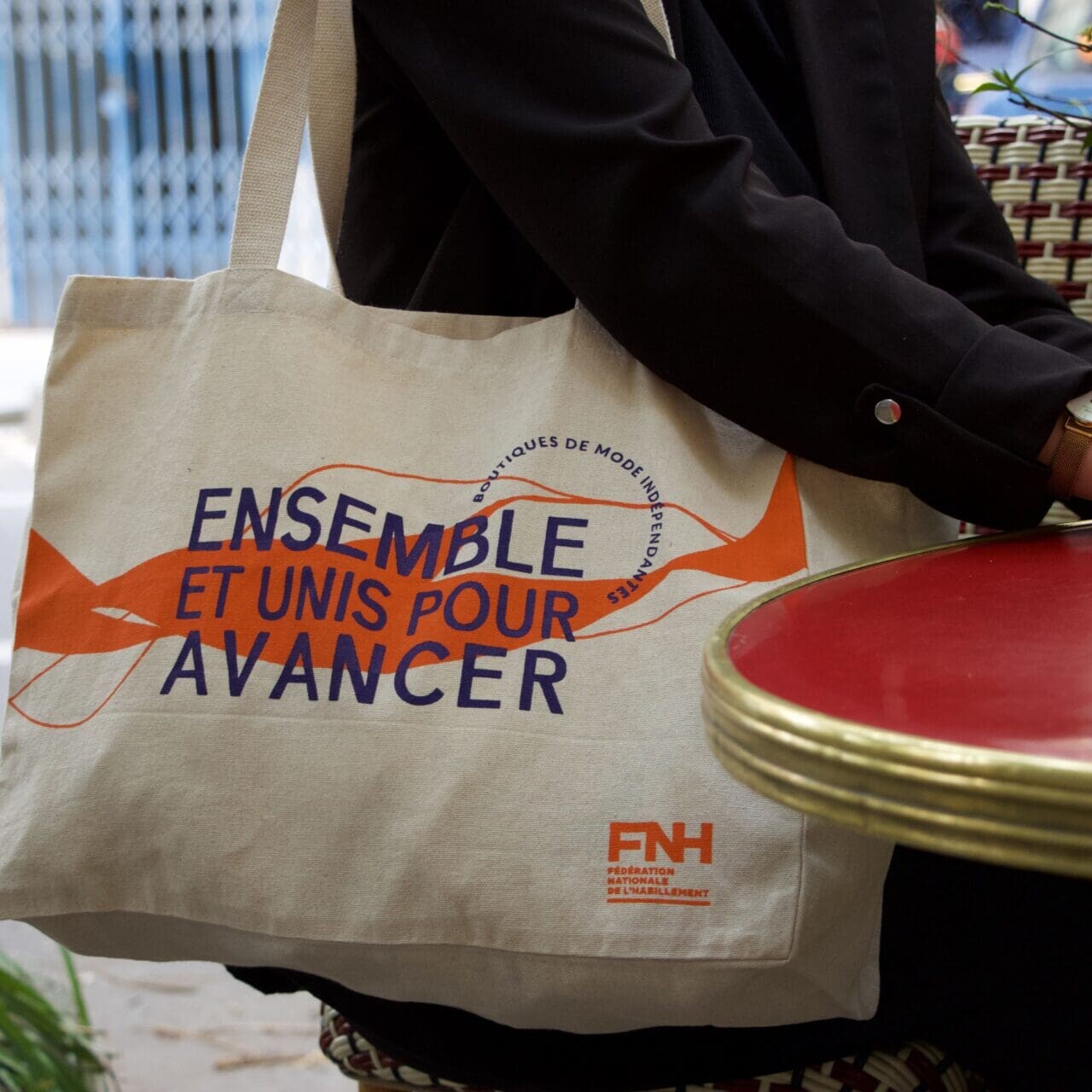 Personne assise à la terrasse d’un café avec un sac fourre-tout beige sur les genoux. Le sac porte un texte orange et violet en français indiquant « Ensemble et unis pour avancer », ainsi qu'un logo orange étiqueté « FNH.