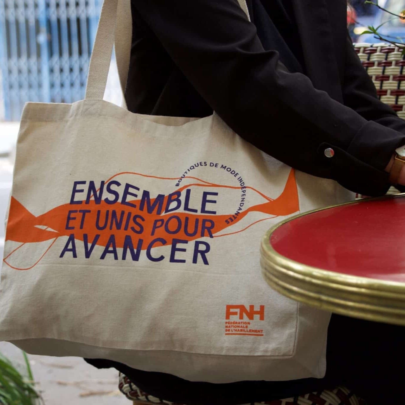 Personne assise à la terrasse d’un café avec un sac fourre-tout beige sur les genoux. Le sac porte un texte orange et violet en français indiquant « Ensemble et unis pour avancer », ainsi qu'un logo orange étiqueté « FNH.