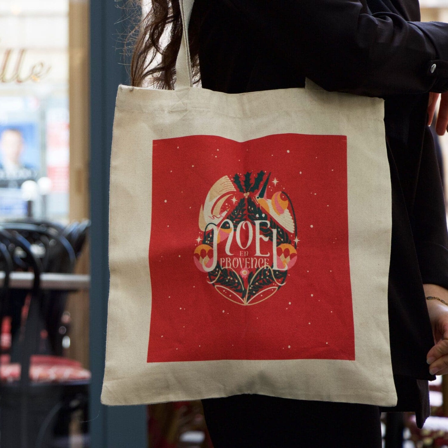 Personne tenant un sac fourre-tout de couleur crème avec un motif carré rouge comportant le texte « NOËL Provençal » et une œuvre d'art sur le thème des fêtes. L'arrière-plan montre un café avec des chaises et des tables.
