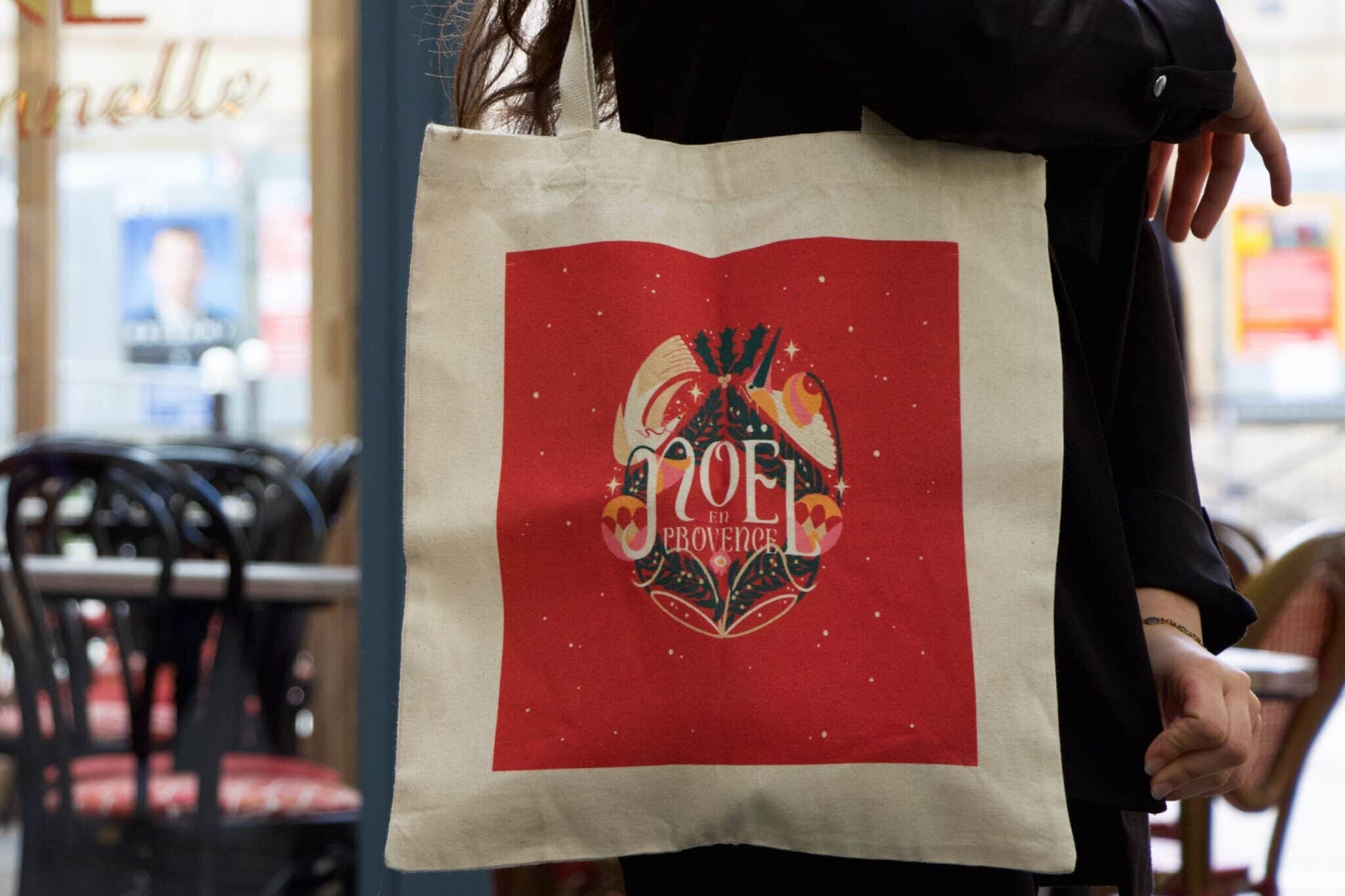 Personne tenant un sac fourre-tout de couleur crème avec un motif carré rouge comportant le texte « NOËL Provençal » et une œuvre d'art sur le thème des fêtes. L'arrière-plan montre un café avec des chaises et des tables.
